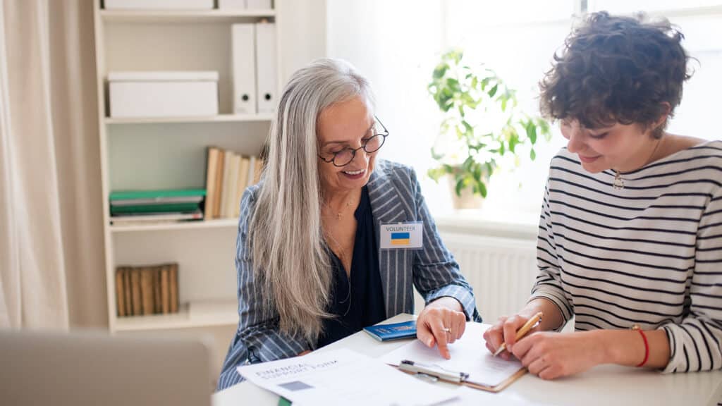 A senior woman volunteer helping Ukrainian woman to fill in forms at asylum centre.